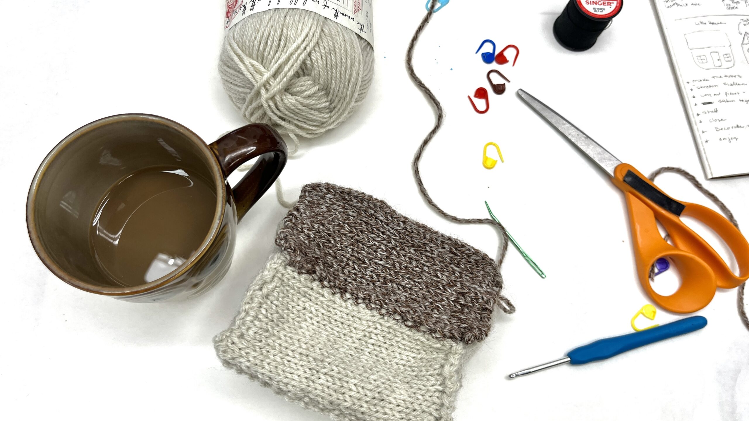Cup of coffee on the left with a soft knitted sculpture of a thatched coastal cottage. Scattered on the table are colorful stitch markers, scissors, crochet hook and a darning needle
